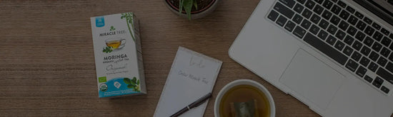 A wooden desk with a laptop, a cup of tea with a teabag, a notepad with handwritten text, and a box of Moringa organic tea. A small potted plant is in the top center of the image.
