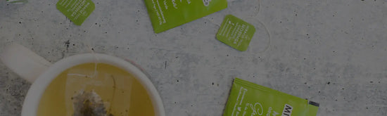 A mug of tea with a tea bag floating inside is on a textured surface. Several green tea bag wrappers and tags are scattered around the mug.