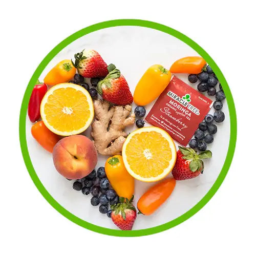 Heart-shaped arrangement of fruits and veggies, including strawberries, orange halves, blueberries, peppers, ginger, and a peach, surrounding a packet labeled Miracle Tree Moringa Superfood. A green circle frames the composition.