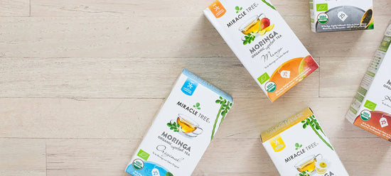 Boxes of Miracle Tree organic moringa tea in various flavors are scattered on a light wooden surface. The visible flavors include Original, Mint, and Mango. Each box features colorful illustrations and labels indicating theyre USDA organic certified.
